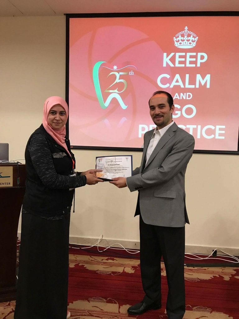 A woman in a pink hijab and a man in a grey suit are exchanging a certificate during a presentation event, with a backdrop that reads 'KEEP CALM AND GO PRACTICE'.