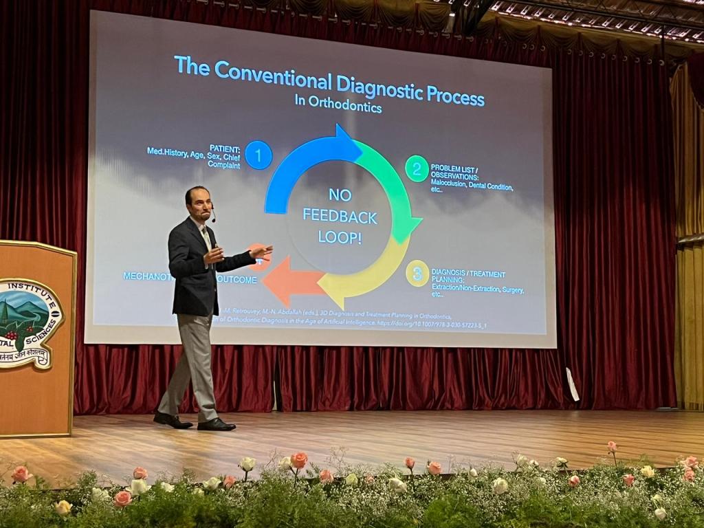 A presenter discussing the conventional diagnostic process in orthodontics, with a slide illustrating the steps involved. The setting features a stage with floral decorations and a logo in the background.
